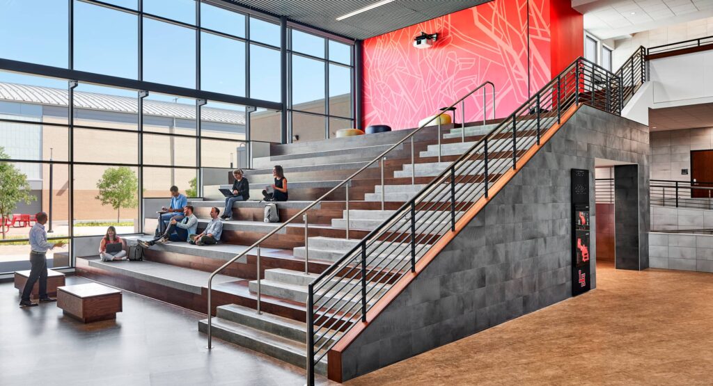 edu-22-H Modern atrium with tiered seating and students engaging in study, large windows, bright red wall art, and railings for safety. | Sky Rye Design Modern atrium with tiered seating and students engaging in study, large windows, bright red wall art, and railings for safety.