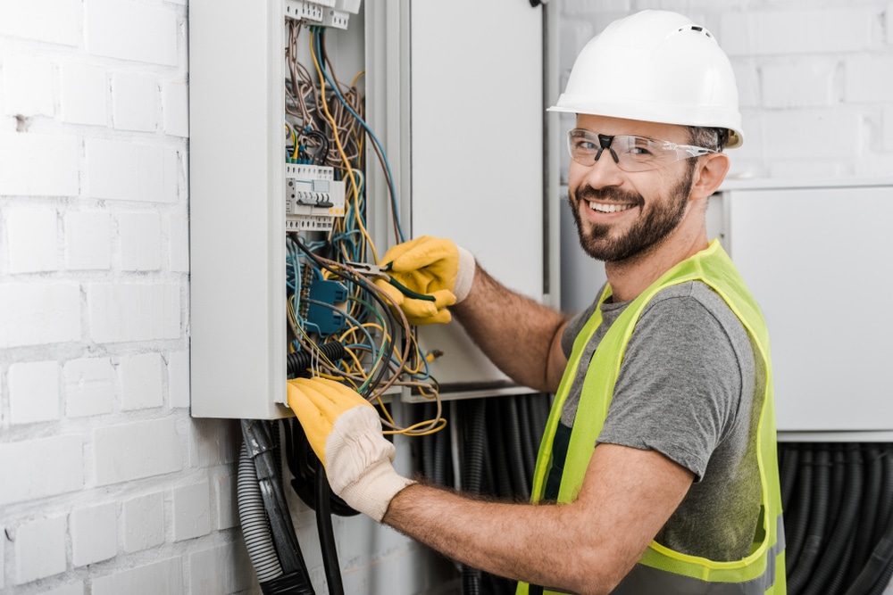 Smiling electrician in safety gear working on an electrical panel. Wearing a hard hat and gloves, checking wires in a control box.