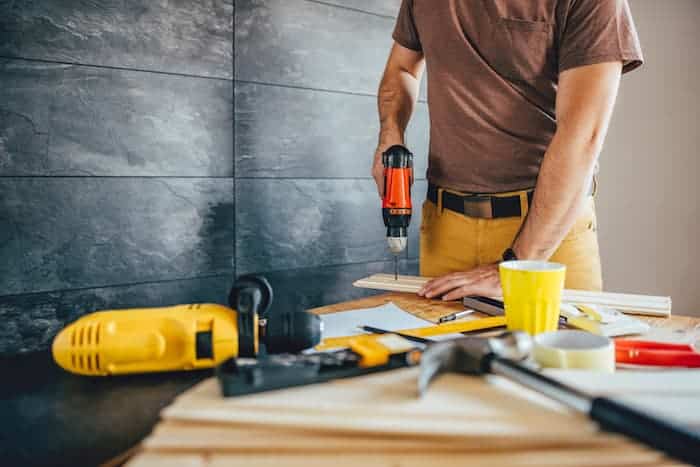 Man using an electric drill on wood with various tools on the table in a workshop setting.