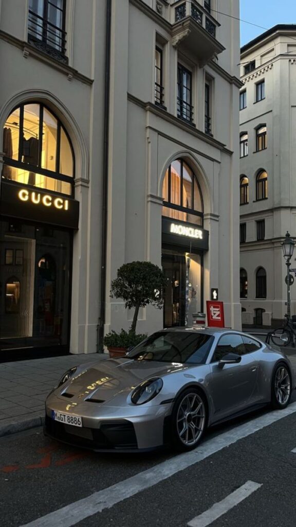 Silver sports car parked in front of Gucci and Moncler stores on a stylish city street at dusk.