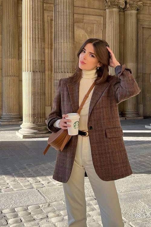 Woman in brown blazer and white turtleneck holding coffee cup, standing on a cobblestone street with historic columns in the background.