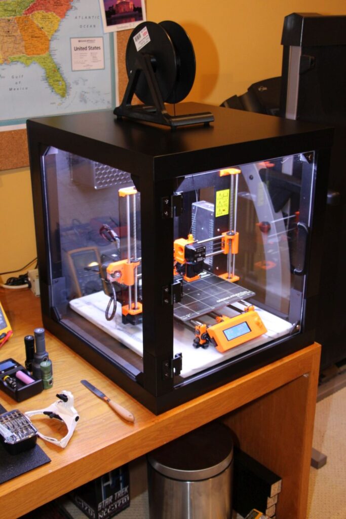 3D printer in a transparent enclosure on a wooden desk, with tools and a map in the background.