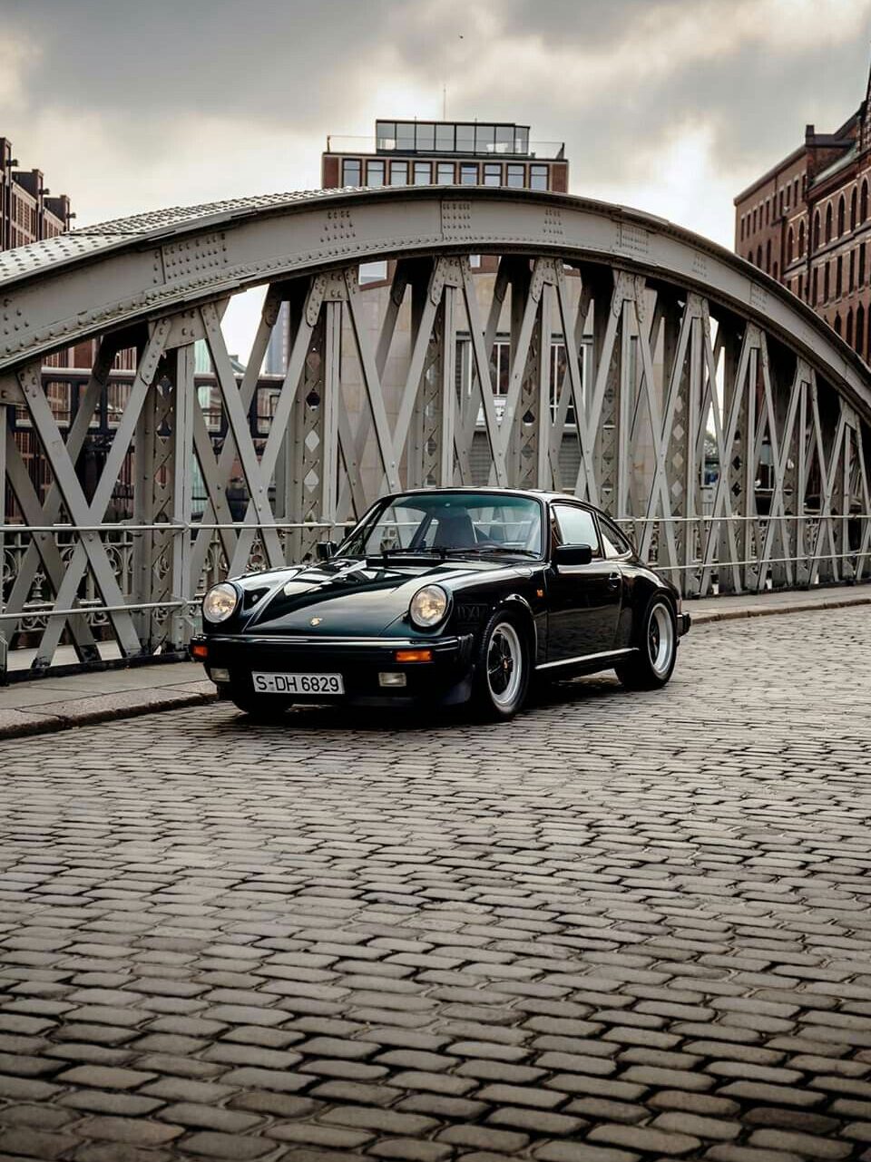 f59d7f1c300f78178debbec139c1f951 Vintage black car on a cobblestone bridge with a dramatic urban background and a bird flying above. | Sky Rye Design Vintage black car on a cobblestone bridge with a dramatic urban background and a bird flying above.