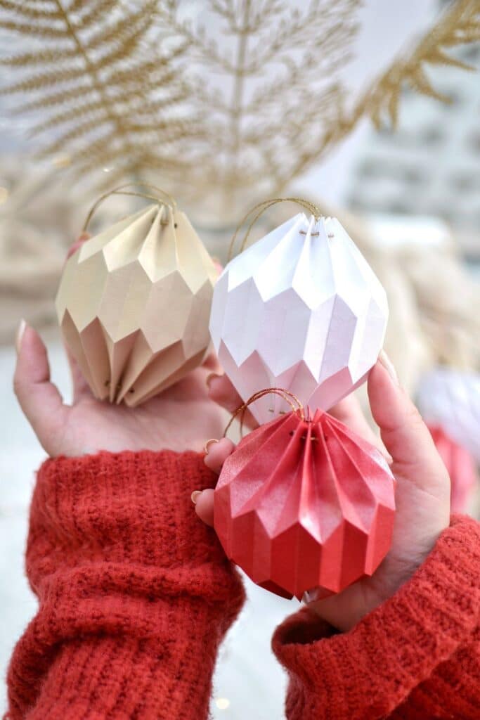 Person holding handmade origami Christmas ornaments in beige, white, and red with golden strings, wearing a red sweater.