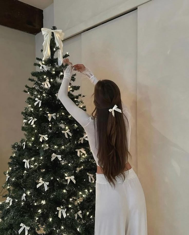 Woman decorating a Christmas tree with white bows and ribbon in a cozy room.