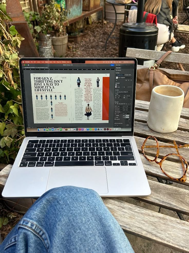 Open laptop on outdoor table, displaying article on thrifting as a lifestyle. Coffee, glasses, and denim jeans visible in relaxed setup.
