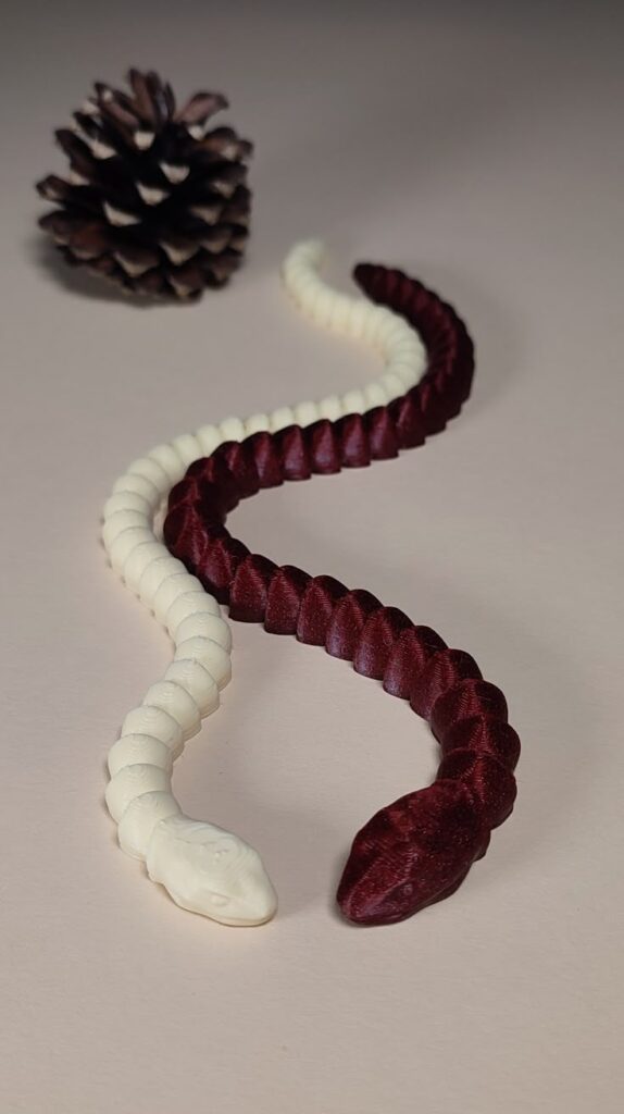Two intertwined red and white 3D-printed snakes near a pine cone on a beige background.