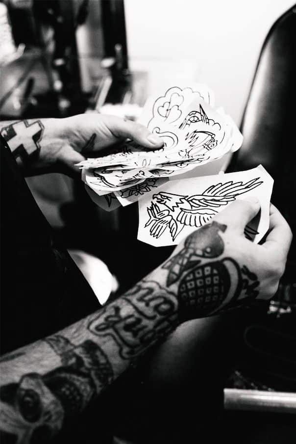 fcf5e88f415b24cc2ea1c5c7a9ff8c25 Tattoo artist holding black and white tattoo designs, showcasing bird and heart motifs, with tattooed arms visible. | Sky Rye Design Tattoo artist holding black and white tattoo designs, showcasing bird and heart motifs, with tattooed arms visible.