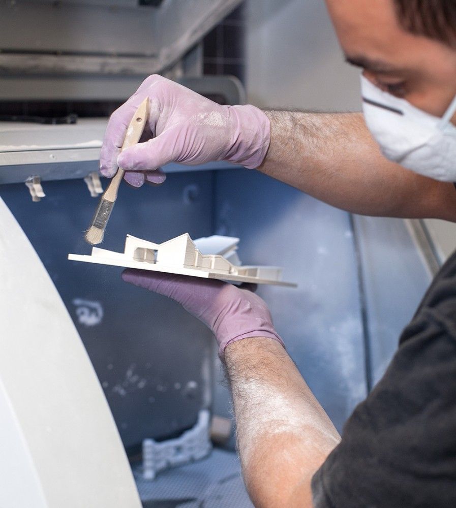 fe586849242a0a5fb2d2d7e67a1fe00a Technician in purple gloves and mask refining a 3D printed object with a brush in a workshop setting. | Sky Rye Design Technician in purple gloves and mask refining a 3D printed object with a brush in a workshop setting.