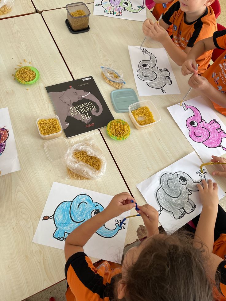 Kids coloring elephant drawings at a table, surrounded by colored pencils, pasta, and a book. Creative activity in a classroom setting.