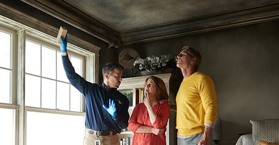 Home inspector wearing gloves examines a smoke-damaged ceiling while explaining findings to concerned homeowners in a dimly lit room.