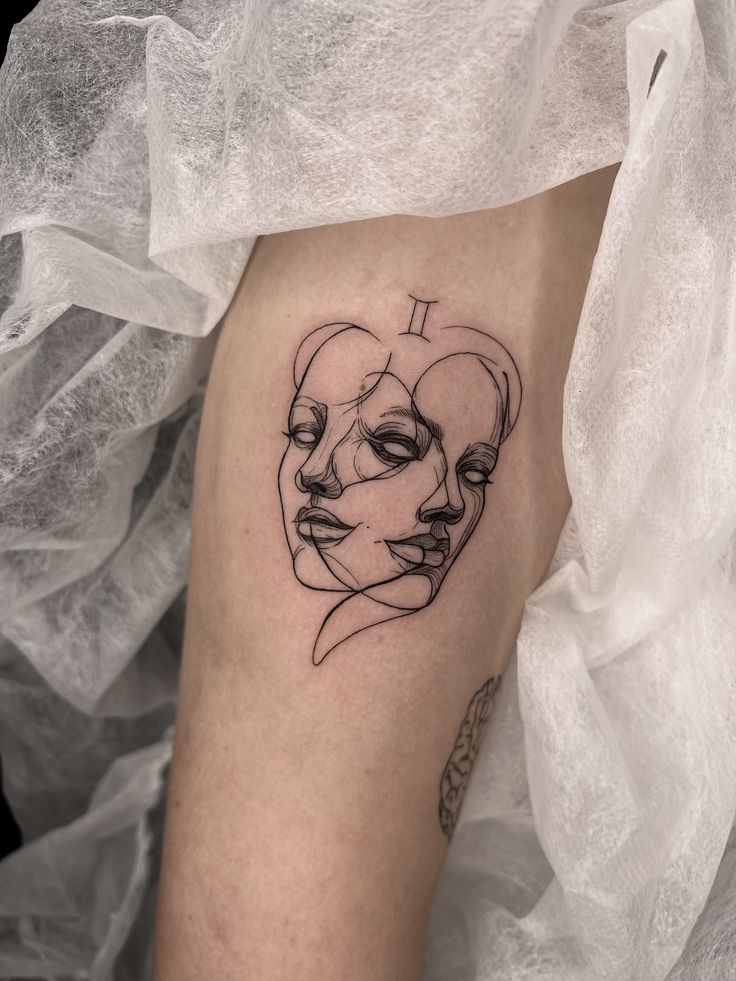 Abstract Gemini tattoo with two overlapping faces and zodiac symbol on arm, set against a textured white fabric background.