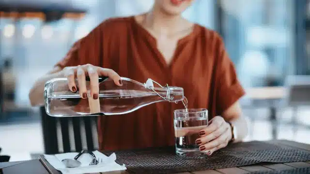 Person pouring water from a glass bottle into a cup on a dining table, emphasizing hydration and sustainable living.
