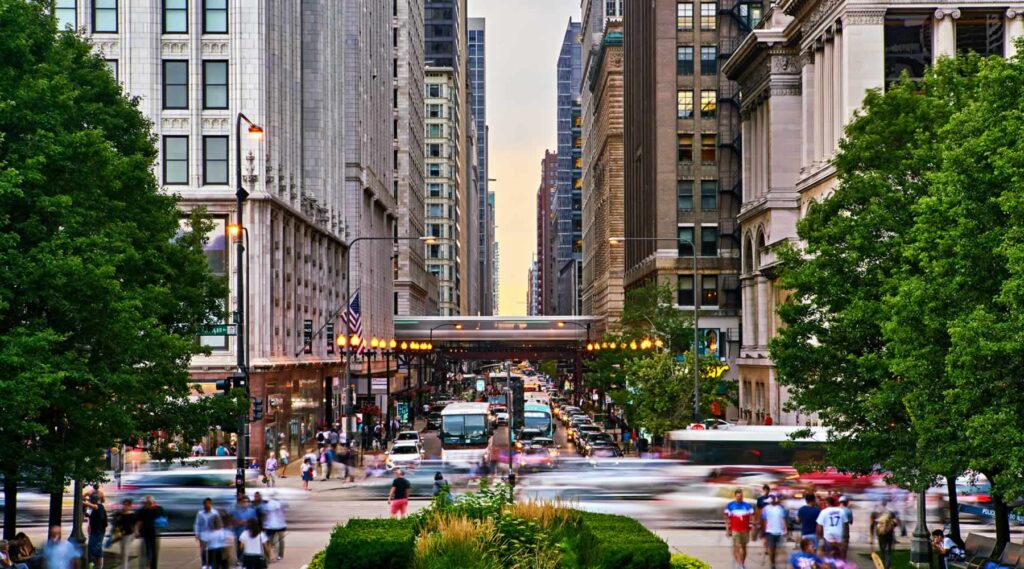 Busy urban street in a vibrant city lined with tall buildings, bustling with people, cars, and a train passing overhead at twilight.