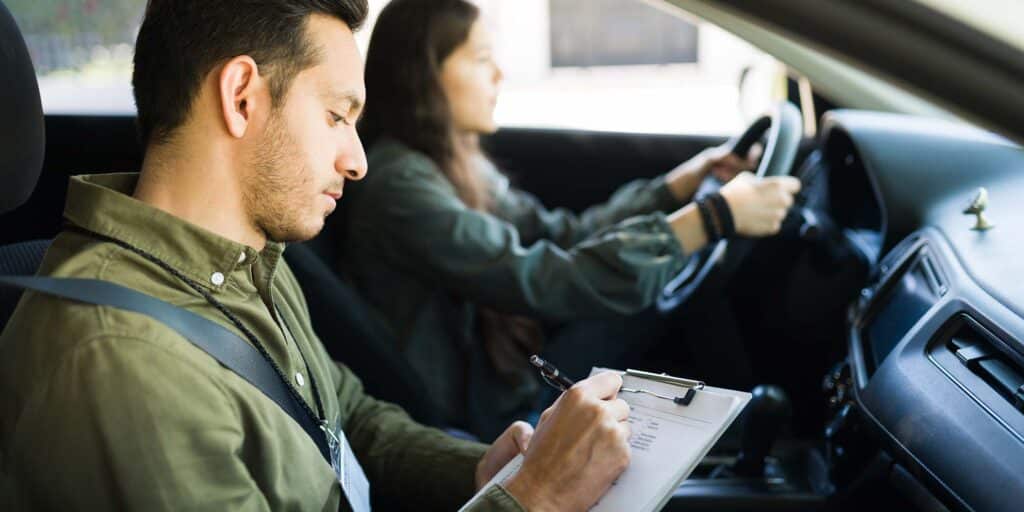 Driving test in progress with a student driver at the wheel and an instructor taking notes inside a car.