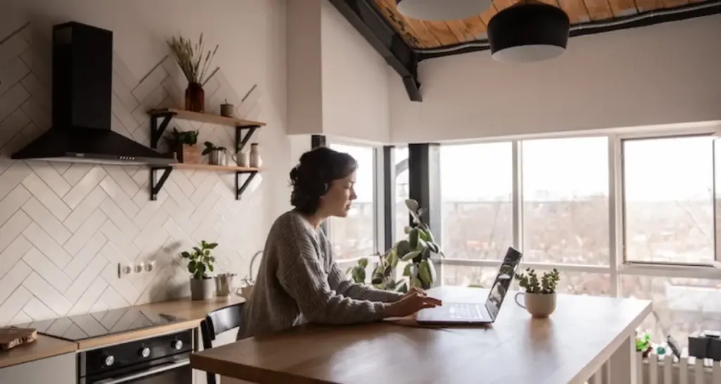 Woman working on a laptop in a modern kitchen with natural light and plants, creating a cozy home office atmosphere.