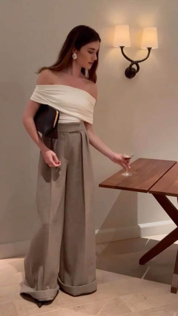 iliridakrasniqi Woman in elegant off-shoulder top and wide-leg trousers holding a clutch and drink in a warmly lit room with wall sconce. | Sky Rye Design Woman in elegant off-shoulder top and wide-leg trousers holding a clutch and drink in a warmly lit room with wall sconce.