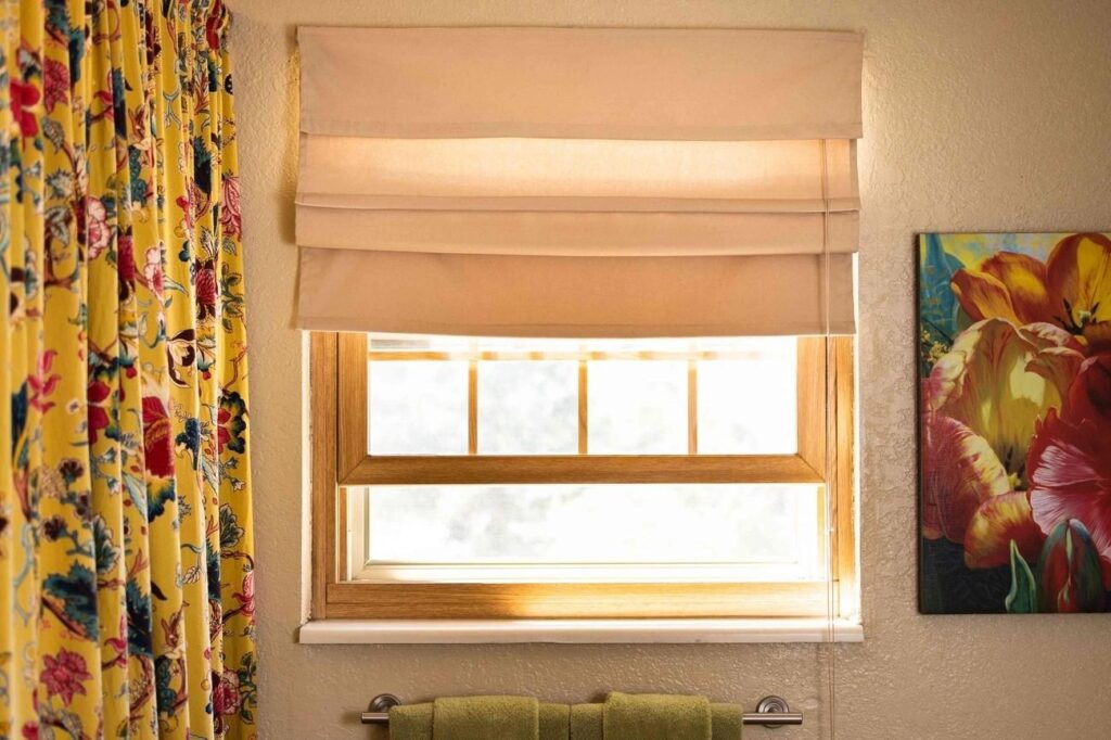 Beige Roman shade on a sunny window, framed by floral curtains and colorful wall art, adding brightness to a cozy interior space.