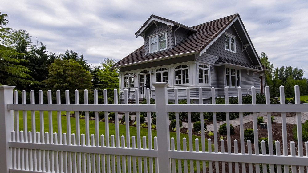 image Charming suburban house with a wooden white fence, lush garden, and cloudy sky backdrop, showcasing a perfect family home. | Sky Rye Design Charming suburban house with a wooden white fence, lush garden, and cloudy sky backdrop, showcasing a perfect family home.