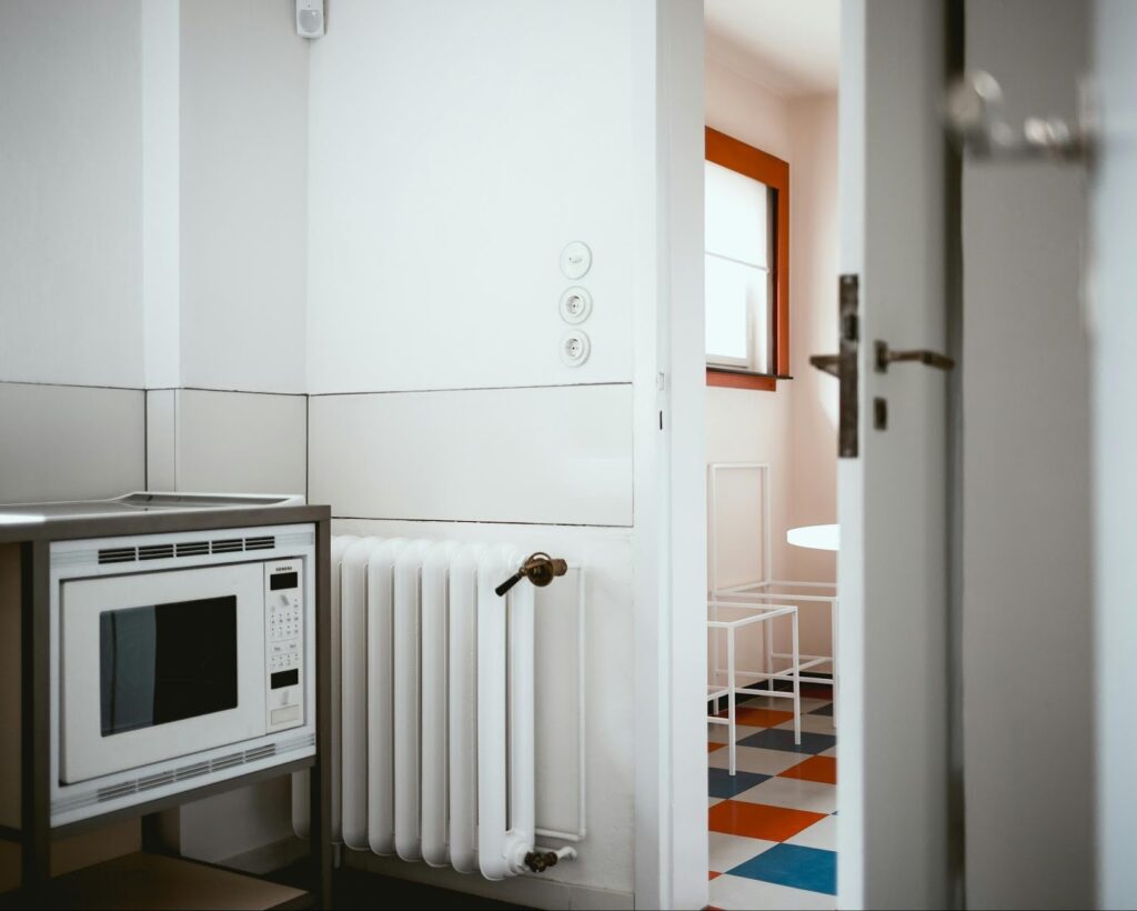 Modern kitchen corner with microwave, radiator, and a glimpse of colorful tiled flooring, viewed through an open door.