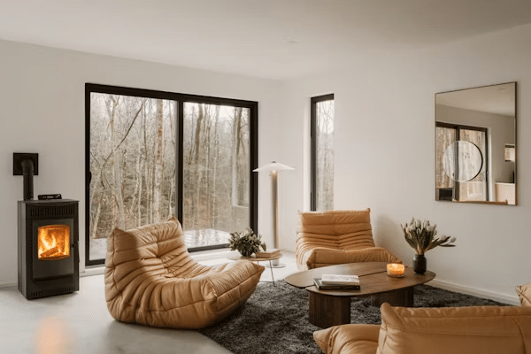 Cozy living room with tan lounge chairs, a modern wood stove, large windows with forest views, and a contemporary coffee table.