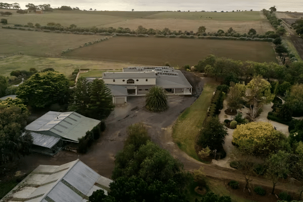 Aerial view of a rural property with a large house surrounded by trees, farmland, and gardens under a clear sky.