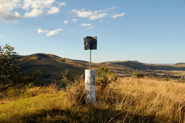 Rural landscape with a weathered, white pillar and signpost on a grassy hill, overlooking rolling hills and a small town under a clear blue sky.