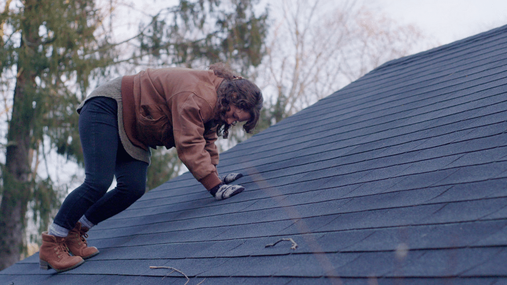 image Person climbing a shingled roof in a jacket and gloves, surrounded by trees in the background. | Sky Rye Design Person climbing a shingled roof in a jacket and gloves, surrounded by trees in the background.