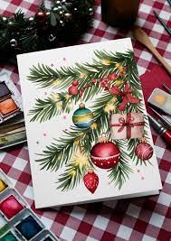 Festive Christmas card with painted evergreen branch, colorful ornaments, gift, and ribbon on checkered tablecloth beside watercolor paints.
