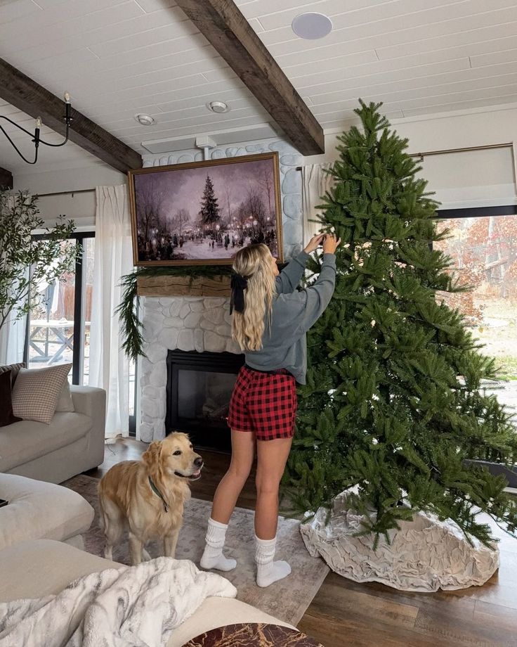 Woman decorating a Christmas tree with her dog nearby in a cozy living room with a fireplace.