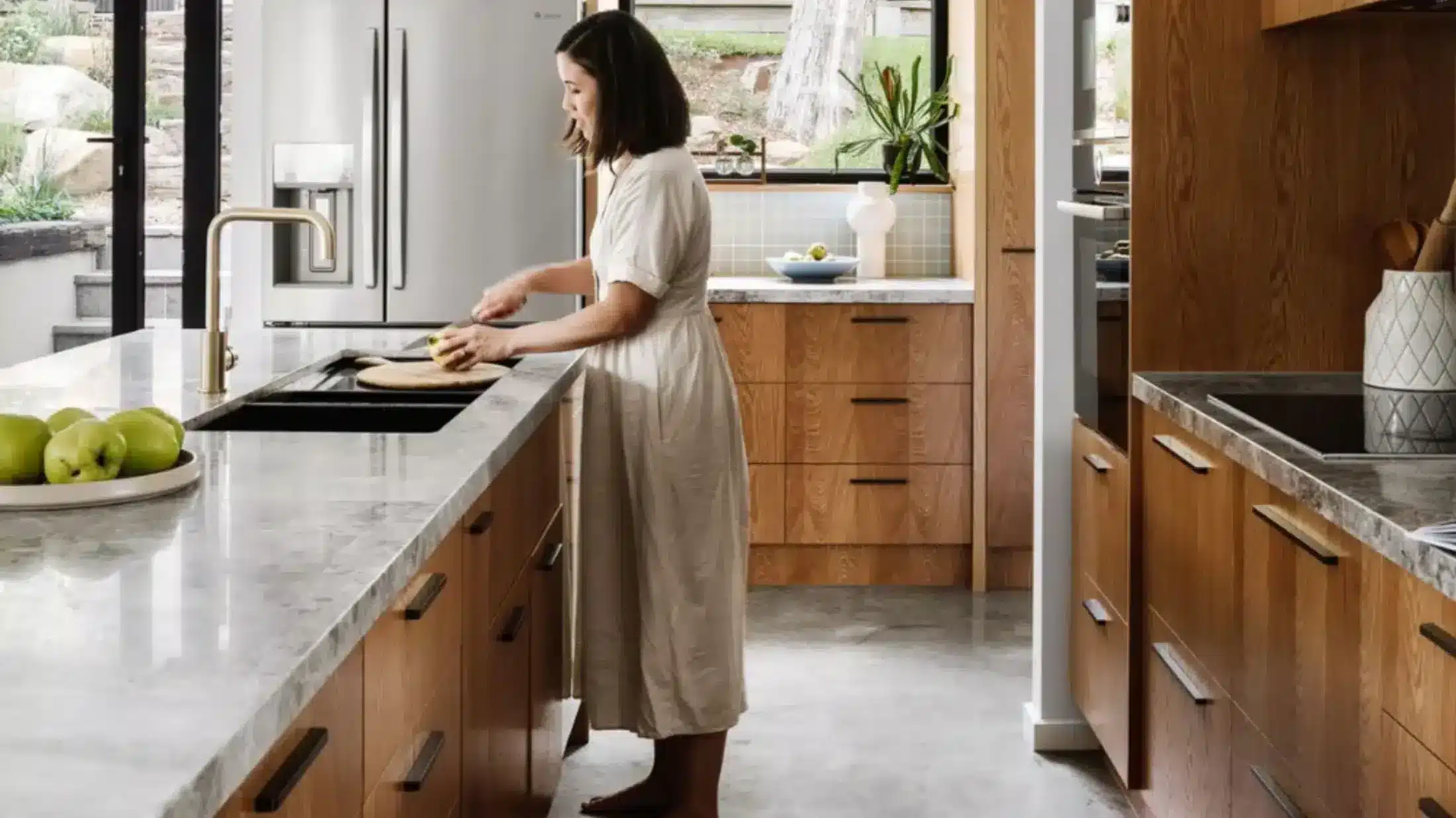 Woman slicing an apple on a wooden countertop in a modern kitchen with wooden cabinets and a view of the garden outside.