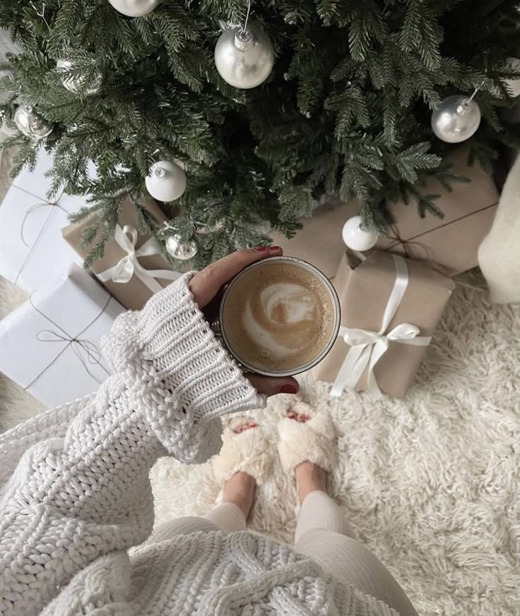 Cozy holiday scene with hot coffee, a Christmas tree, presents, and a person in a knit sweater and slippers.