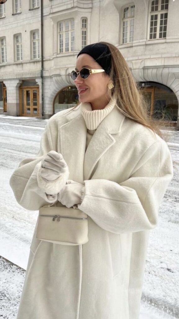 Woman in elegant white winter coat with sunglasses and gloves standing in snowy city street.