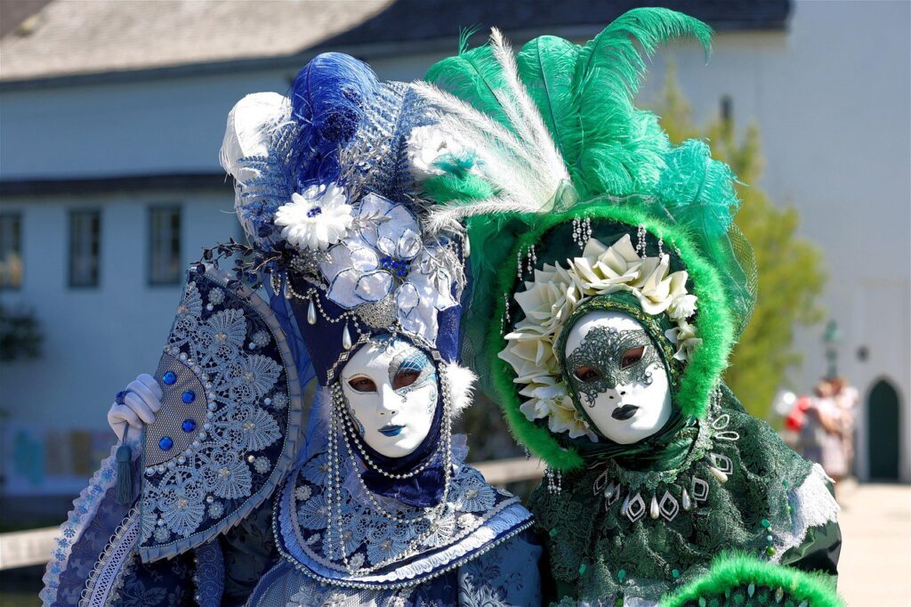 photo_5213365161295547147_y Two people in ornate blue and green Venetian masks adorned with flowers and feathers at a costume event. | Sky Rye Design Two people in ornate blue and green Venetian masks adorned with flowers and feathers at a costume event.