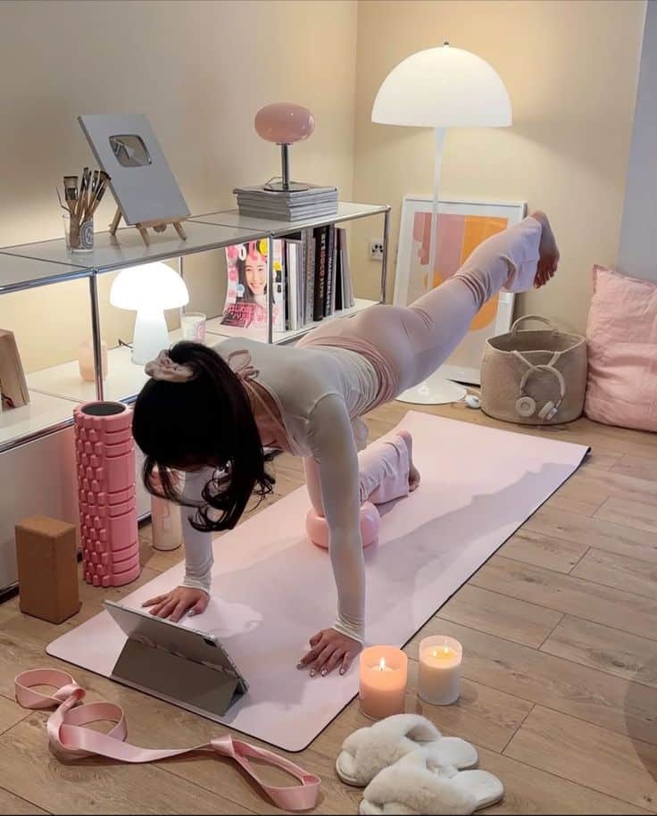 Woman practicing yoga on a mat with a tablet, surrounded by candles and cozy decor in a home setting.