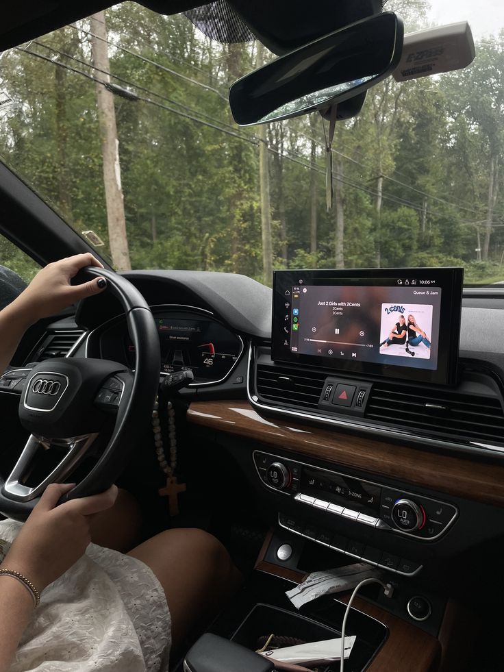 Driver holds steering wheel in an Audi, with music playing on the car's touchscreen against a backdrop of lush greenery outside.