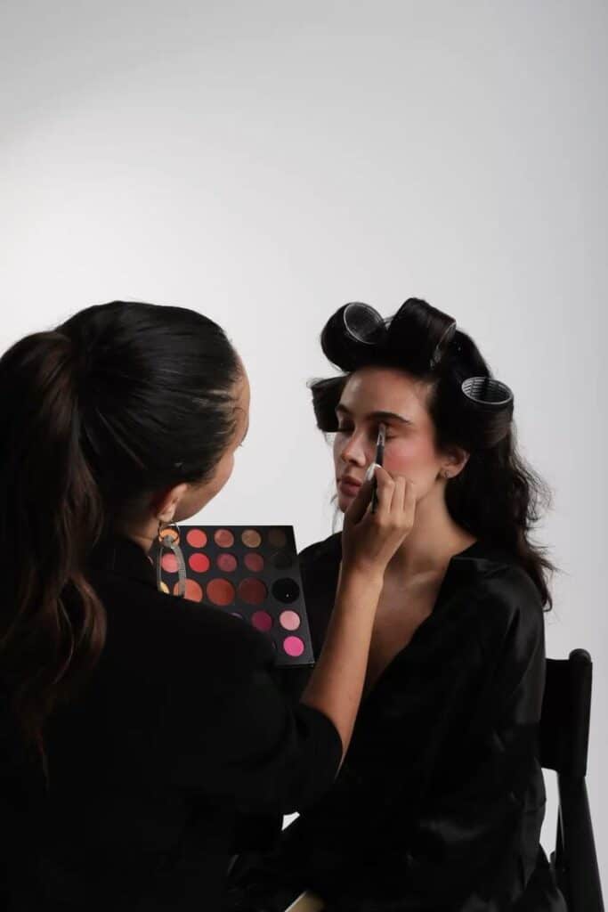 Makeup artist applying eyeshadow to a client with hair rollers in a bright studio setting.