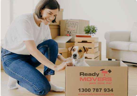 Smiling woman packing with Ready Movers box, cute dog inside, preparing for a move. Boxes and sofa in background.