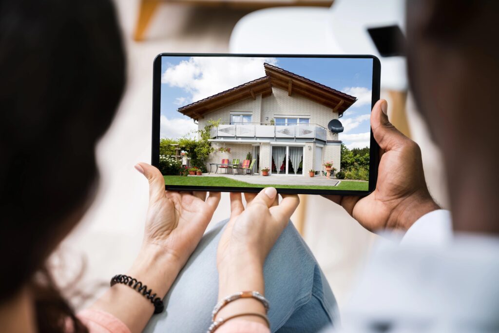 Two people holding a tablet, viewing a digital image of a modern house with a garden, symbolizing real estate and home browsing.