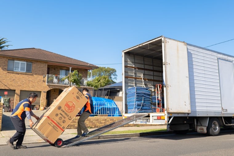 Movers unloading a large box from a truck using a dolly on a residential street with brick house and clear sky in background.