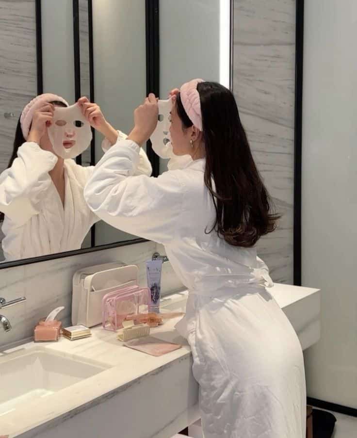 Woman applying a sheet mask in a bathroom, wearing a robe and headband, with skincare products on the marble counter.