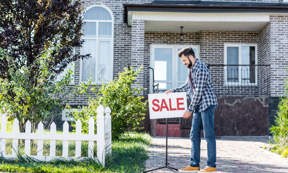 Man placing a Sale sign outside a brick house with a white picket fence.
