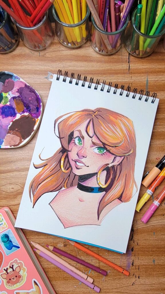 Spiral sketchbook with colorful portrait of a woman, surrounded by colored pencils and art supplies on a wooden desk.