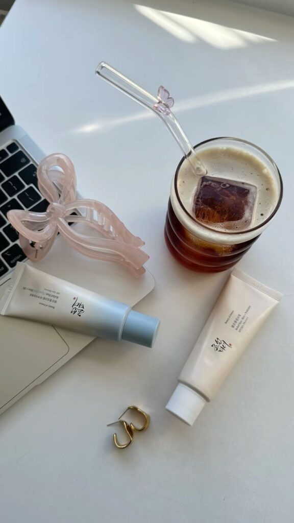 Skincare tubes on laptop, iced coffee with straw, hair clip, and gold earrings on a white desk.
