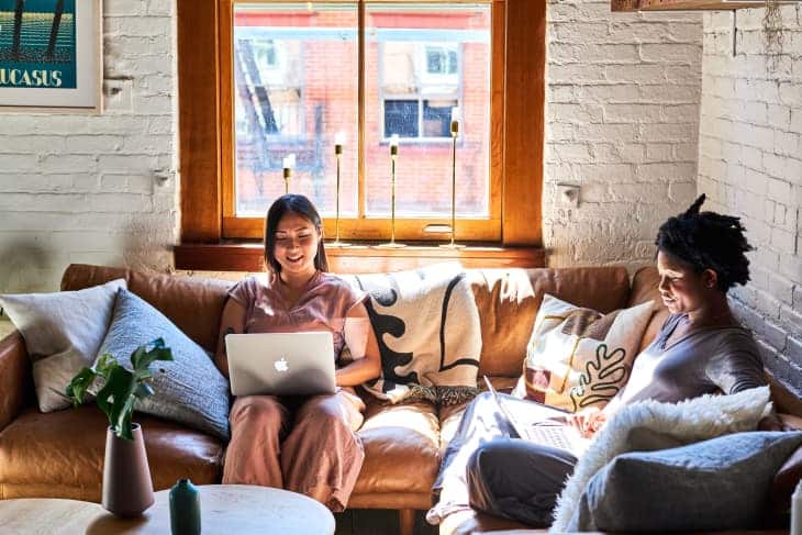 Two people sitting on a cozy couch, using laptops, enjoying natural sunlight through a large window in a stylish living room.