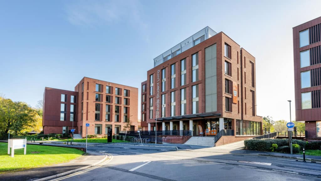 student-accommodation-guildford-bankside-entrance-external Modern student accommodation building with red brick facade and large windows, set on a sunny day with clear skies and greenery. | Sky Rye Design Modern student accommodation building with red brick facade and large windows, set on a sunny day with clear skies and greenery.