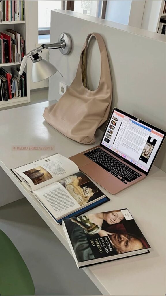 Modern workspace with an open book, pink laptop, and beige bag on a desk. Shelves with books and a lamp are in the background.