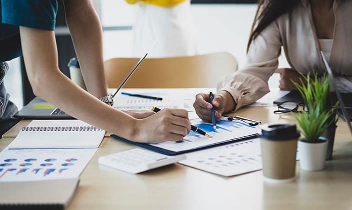 Collaborative team analyzing financial charts and data at a desk with coffee, plants, and supplies, highlighting business strategy discussions.