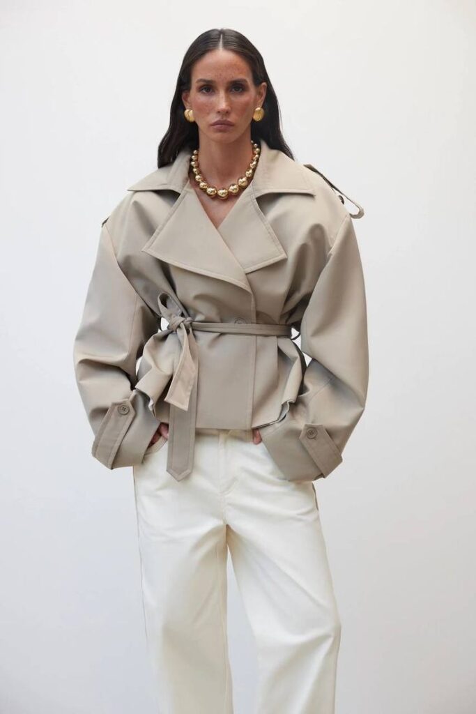 Woman in beige trench coat with gold jewelry and white pants, posing confidently against a plain background.