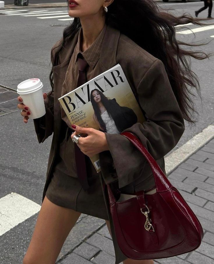 Woman in stylish brown suit with red bag, holds magazine and coffee, walking on city street.